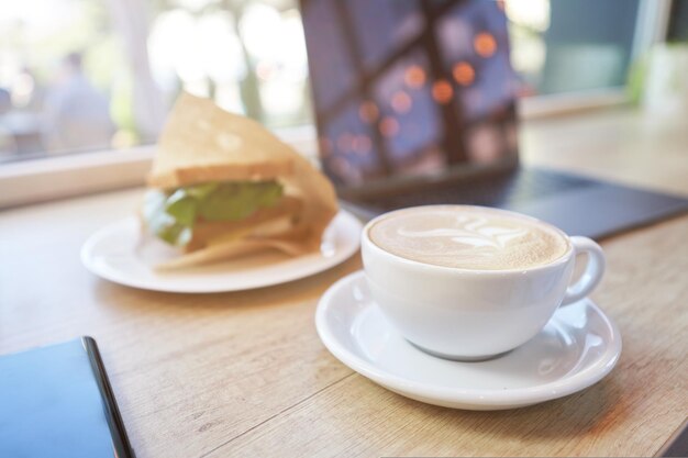 Travailler à distance dans un café en gros plan d'une tasse de café sandwich au cappuccino et d'un ordinateur portable sur bois
