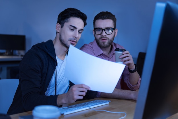 Travailler ensemble. Beaux programmeurs masculins intelligents assis ensemble et regardant la feuille de papier tout en discutant de leur projet