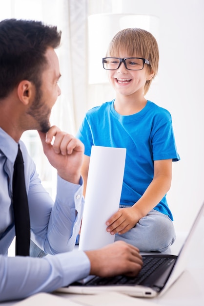 Travailler ensemble est amusant. Joyeux jeune homme en chemise et cravate travaillant sur un ordinateur portable et regardant son fils souriant assis près de lui et tenant des documents