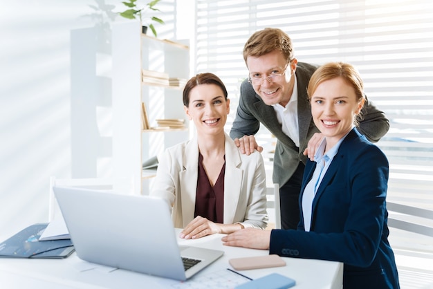 Travailler en équipe. Satisfait joyeux trois collègues souriant tout en regardant la caméra et l'homme plaçant les mains sur les épaules des femmes