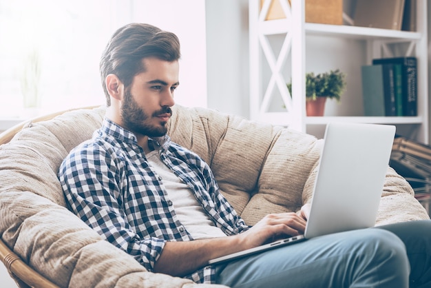 Travailler à la maison. Beau jeune homme travaillant sur ordinateur portable assis dans une grande chaise confortable à la maison