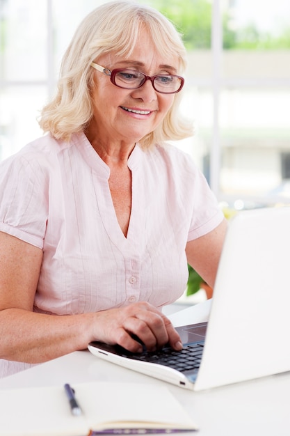 Travailler à la maison. Heureuse femme âgée travaillant sur l'ordinateur portable et souriante assise à la table