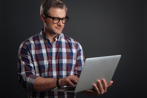 Travailler de n'importe où. Beau mec motivé concentré écrivant un e-mail tout en utilisant son ordinateur portable et portant les lunettes