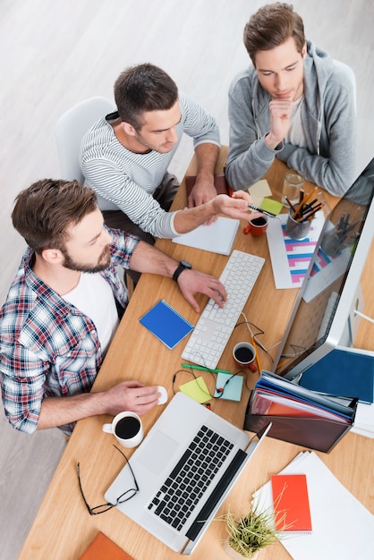 Travailler sur un nouveau projet. Vue de dessus de trois jeunes hommes concentrés travaillant ensemble assis sur le lieu de travail