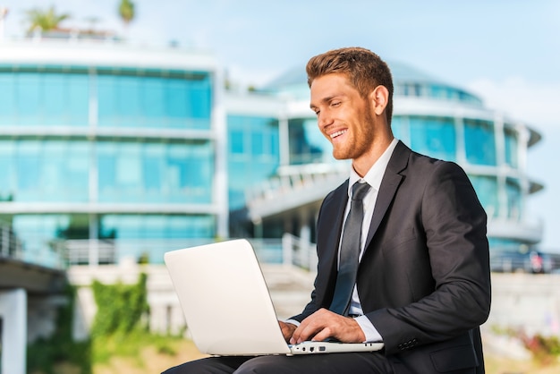 Travailler avec plaisir. Beau jeune homme en tenue de soirée travaillant sur un ordinateur portable et souriant assis à l'extérieur et contre la structure du bâtiment