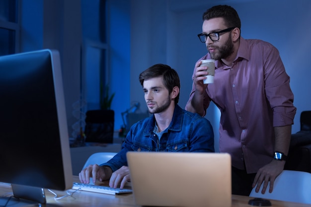 Travailler avec la technologie moderne. Joli programmeur masculin attrayant tapant sur le clavier et regardant l'écran de l'ordinateur tout en travaillant avec son collègue