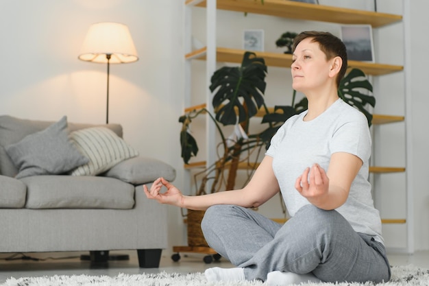 Travailler avec votre corps Toute la longueur d'une belle femme d'âge moyen en vêtements de sport faisant la pose de lotus tout en étant assise devant la fenêtre à la maison