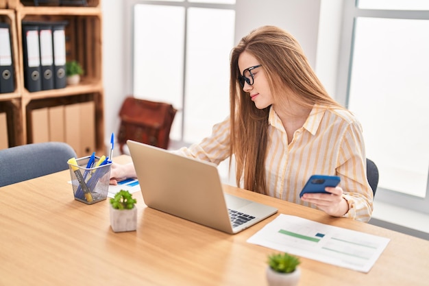 Travailleur d'affaires de jeune femme blonde à l'aide d'un ordinateur portable et d'un smartphone au bureau