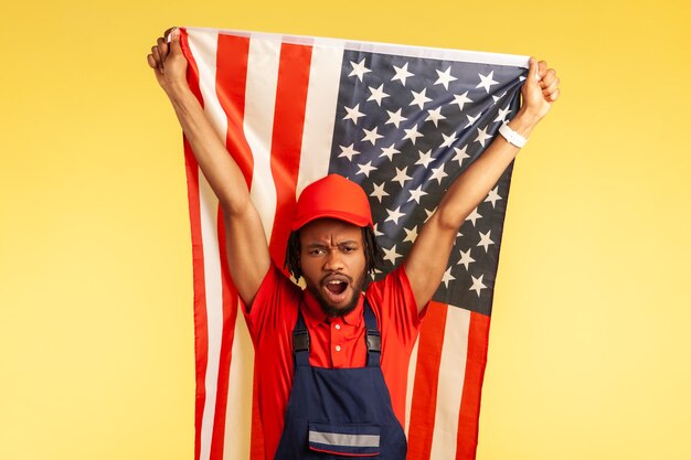 Travailleur afro-américain sérieux en uniforme avec des dreadlocks tenant le drapeau américain et criant, célébrant la liberté et l'indépendance. Tourné en studio intérieur isolé sur fond jaune