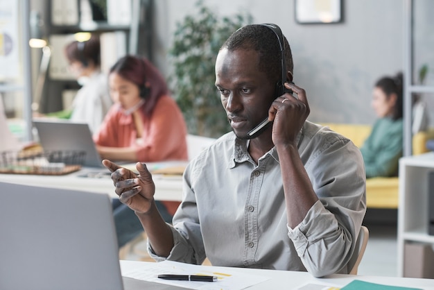 Travailleur d'appel africain dans des écouteurs parlant au téléphone avec des clients à sa table avec un ordinateur portable