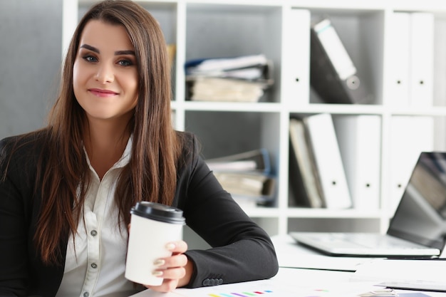 Travailleur attrayant en costume tenant une tasse de café posant sur le lieu de travail au bureau