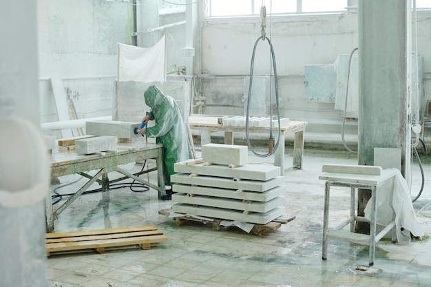 Travailleur en combinaison de protection polissant la pierre avec une meuleuse dans l'atelier de l'usine