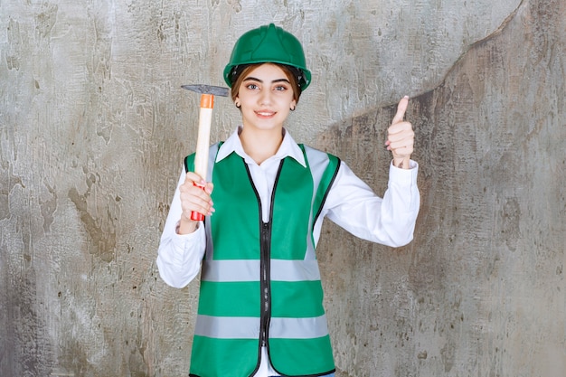 Travailleur de la construction féminin en casque vert tenant un marteau et donnant le pouce vers le haut