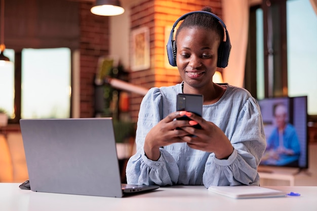 Travailleur à distance afro-américain souriant dans un casque en tapant un message sur son téléphone portable. Femme indépendante regardant des vidéos sur smartphone dans un bureau à domicile moderne avec une belle lumière chaude au coucher du soleil