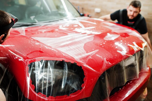 Le travailleur du service automobile a mis un film anti-gravier sur une carrosserie de voiture rouge à l'atelier de détail du véhicule. Protection de voiture avec des films spéciaux.