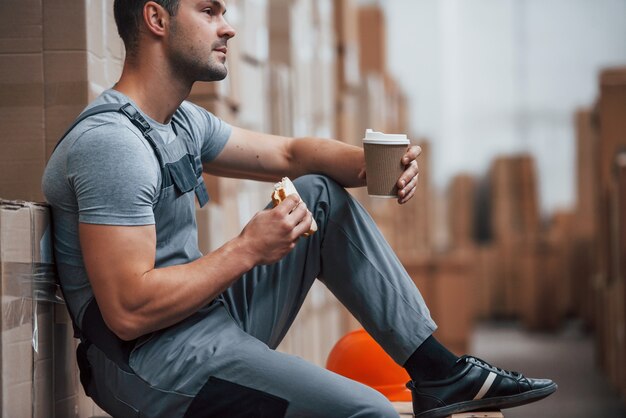 Le travailleur du stockage s'assoit et fait une pause. Mange du sandwitch et boit du café.