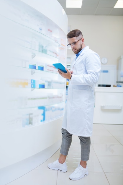 Travailleur élégant. Jeune chimiste concentré regardant une tablette tout en recherchant une liste de médicaments