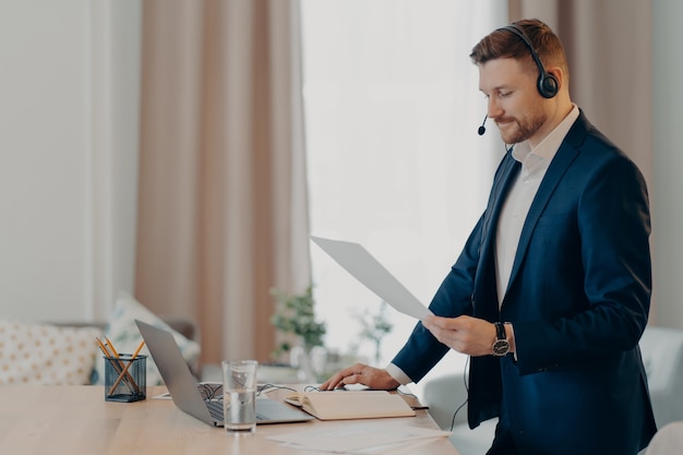 Un travailleur exécutif sérieux donne une présentation du plan d'affaires tient des supports de documents papier près du bureau habillé formellement utilise un ordinateur portable et un casque modernes pour une conférence en ligne. Travail social à distance