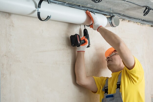 Le travailleur fixe les tuyaux de ventilation au plafond avec un tournevis.