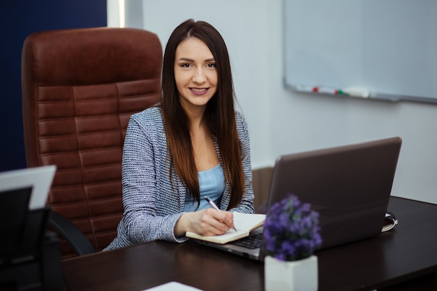 Travailleur gai taper sur un ordinateur portable et regarder ailleurs avec un sourire éclatant. Elle est assise à un bureau dans un bureau. Portrait