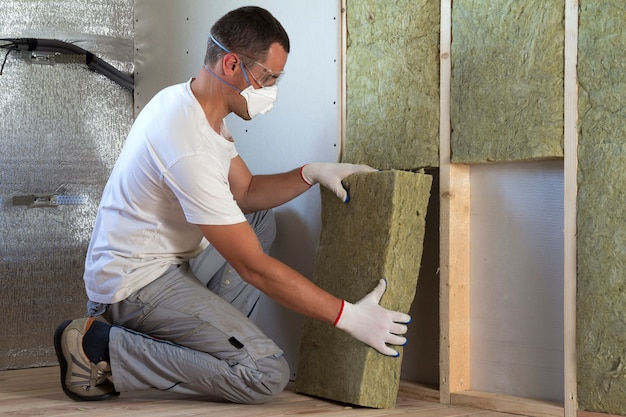 Travailleur en lunettes de protection et respirateur isolant isolant en laine de roche dans un cadre en bois pour les futurs murs de la maison pour la barrière contre le froid. Maison chaleureuse confortable, économie, construction et rénovation