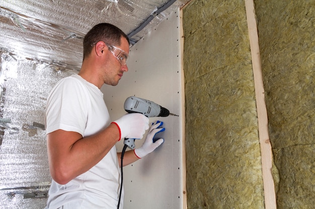 Travailleur en lunettes avec tournevis travaillant sur l'isolation. Cloison sèche sur poutres murales, bâton isolant en laine de roche dans un cadre en bois. Concept d'accueil, d'économie, de construction et de rénovation chaleureux et confortable.