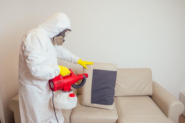 Travailleur de la lutte antiparasitaire en uniforme pulvérisant des pesticides sous le canapé dans le salon