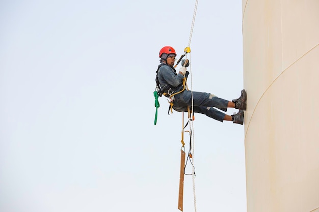 Travailleur masculin accès à la corde industriel travaillant à la hauteur du réservoir d'huile portant un harnais, équipement de sécurité du casque inspection de l'accès par corde de l'épaisseur du réservoir de pétrole et de gaz.