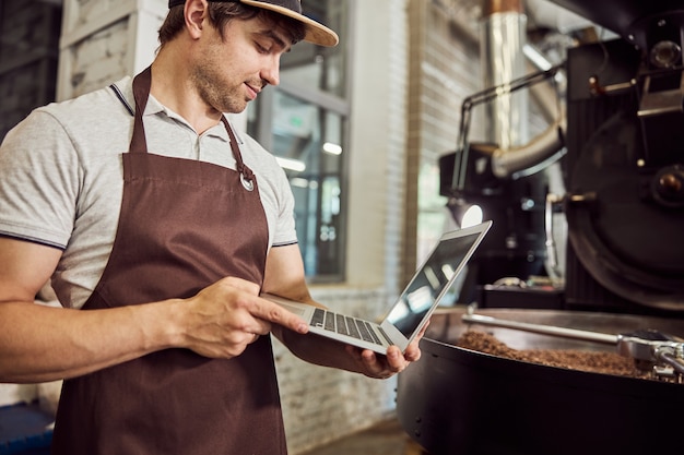 Travailleur masculin attrayant en tablier tenant un ordinateur portable moderne tout en se tenant près d'une machine de torréfaction de café professionnelle