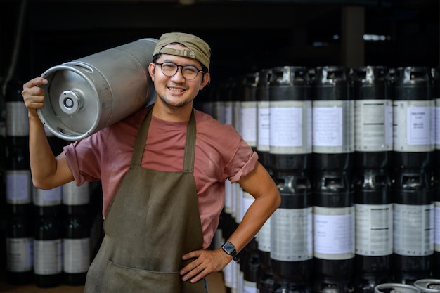 Travailleur masculin avec des fûts de bière en métal dans une brasserie Petite entreprise et l'industrie de la bière Souriant bel homme asiatique dans un tablier tenant un seau en acier sur son épaule dans un entrepôt