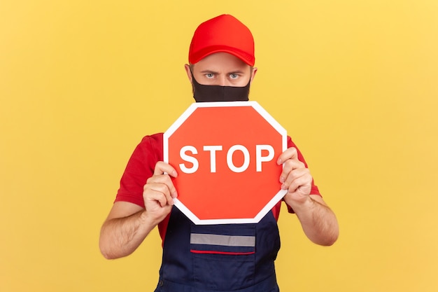 Travailleur masculin en salopette bleue chapeau rouge et tshirt avec masque de protection tenant un panneau de signalisation d'arrêt