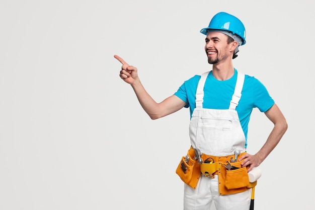 Travailleur masculin souriant confiant dans l'ensemble et casque avec boîte à outils pointant sur un espace vide