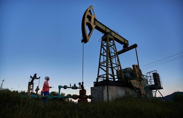 Travailleur masculin tenant un presse-papiers et regardant la prise de pompe à pétrole tout en travaillant dans un champ pétrolifère Homme pétrolier debout près de l'unité de pompage de pétrole sous le ciel bleu Concept d'extraction de pétrole et d'industrie pétrolière