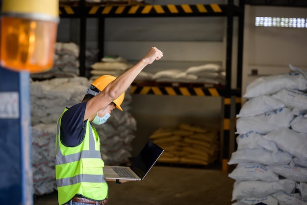 Un travailleur masculin utilisant un ordinateur tablette travaillant dans une usine de produits chimiques industriels ou d'emballage porte un casque orange dans un entrepôt industriel L'arrière-plan est un entrepôt et des marchandises