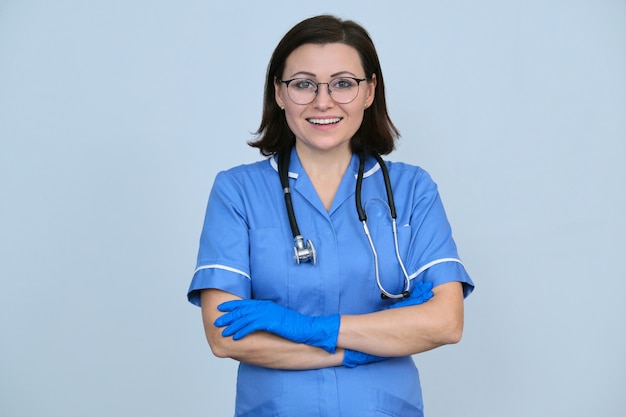 Travailleur médical féminin en uniforme bleu avec stéthoscope et gants, confiant femme professionnelle avec les bras croisés regardant la caméra, fond gris clair