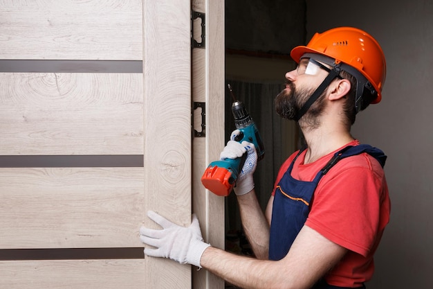 Un travailleur avec une perceuse dans un casque orange installe une porte dans la maison