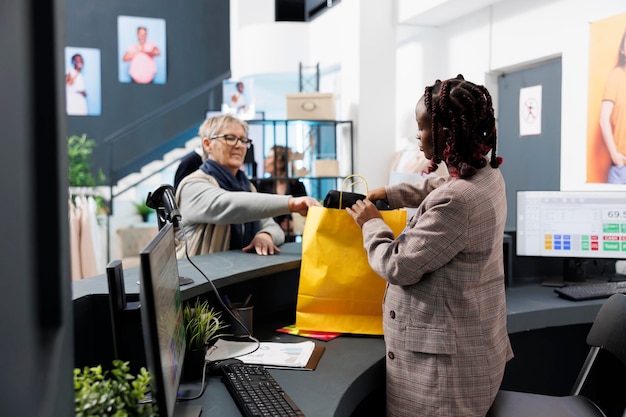 Travailleur de salle d'exposition afro-américain préparant l'achat pour un client âgé dans un magasin de vêtements. Femme accro du shopping achetant des vêtements décontractés à la mode, effectuant un paiement électronique au terminal pos.