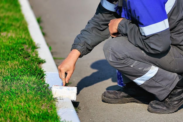 Un travailleur en salopette peint une bordure avec de la peinture blanche un jour d'été Aménagement paysager des services urbains