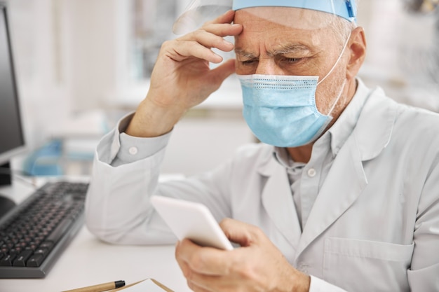 Travailleur de la santé âgé portant un masque de protection et ayant l'air réfléchi tout en vérifiant son téléphone