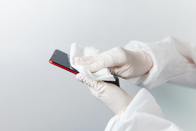 Travailleur de la santé portant des EPI et des gants en latex désinfectant un téléphone portable sur fond blanc