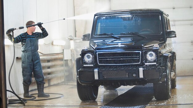 Travailleur en service automobile lave une voiture de luxe dans la mousse par téléobjectif de tuyaux d'eau