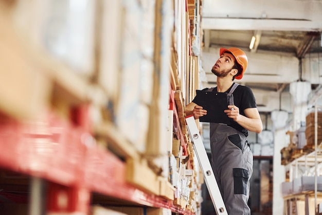 Le travailleur de stockage se tient sur l'échelle en uniforme et le bloc-notes dans les mains et vérifie la production.