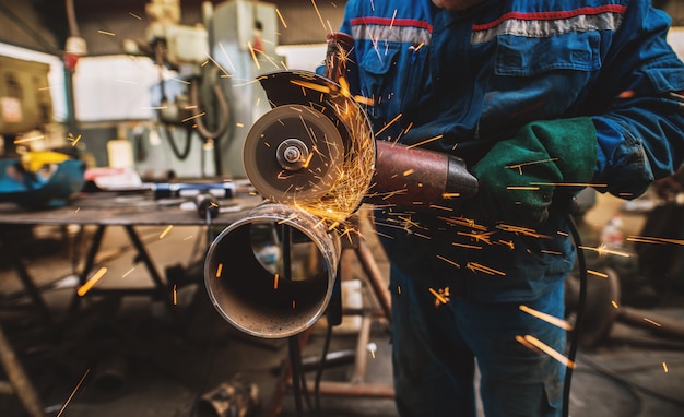 Travailleur en tissu en uniforme de protection de tuyau métallique de coupe sur la table de travail avec une meuleuse électrique dans l'atelier industriel.