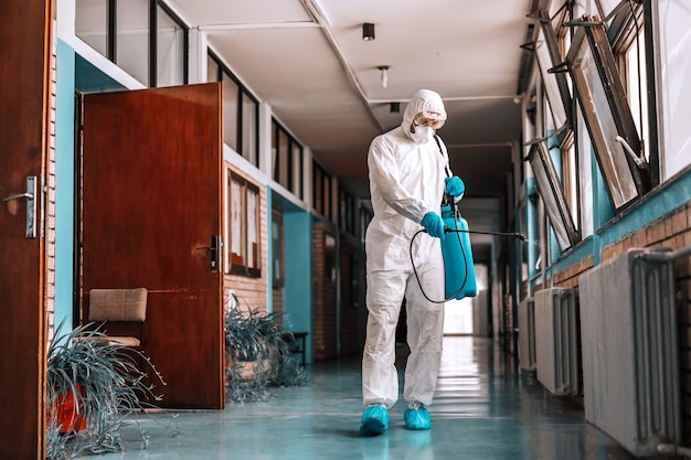 Travailleur sur toute la longueur en uniforme stérile, avec masque facial tenant un pulvérisateur avec désinfectant et pulvérisation dans le couloir de l'école.