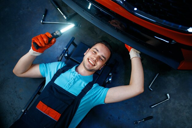 Travailleur en uniforme couché sous le véhicule, station-service de voiture. Contrôle et inspection automobile, diagnostic et réparation professionnels