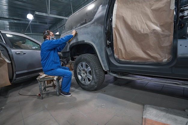 Photo travailleur en vêtements de protection assis près de la voiture et faisant le meulage des carrosseries de voiture avant de peindre et d'apprêter