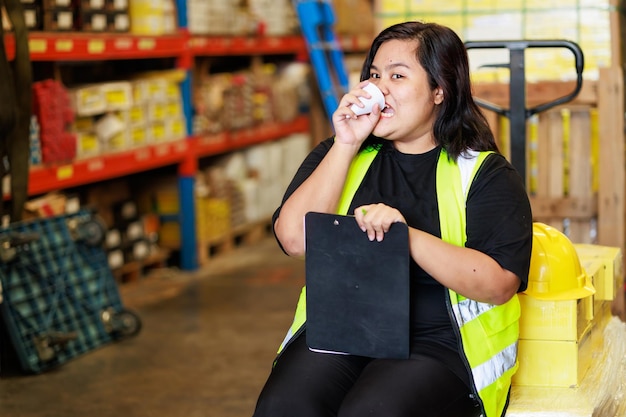 Les travailleurs boivent de l'eau, du café ou du thé pendant la pause Chaude fatigue et épuisement Asiatique grosse femme d'entrepôt obèse en gilet de sécurité et casque de protection à l'usine d'entrepôt industrielle