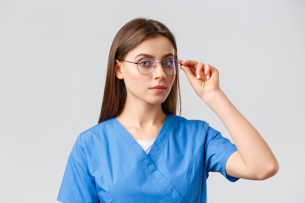 Travailleurs de la santé, médecine, assurance et concept de pandémie de covid-19. Infirmière professionnelle intelligente, médecin ou stagiaire en gommages bleus et lunettes regardant la caméra déterminée, fond gris