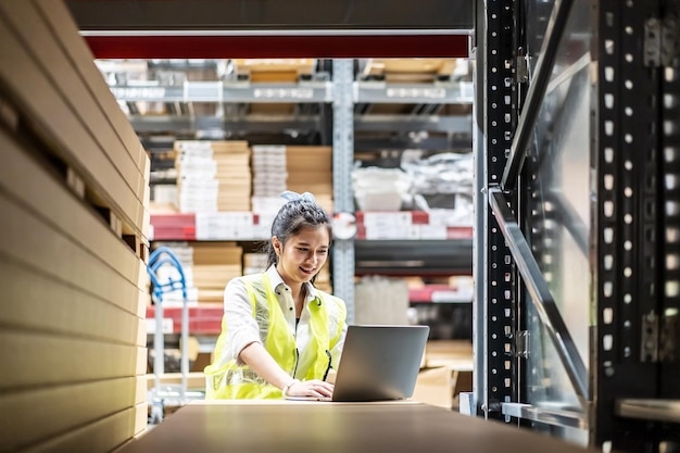 Travailleuse asiatique en gilet de sécurité assise et travaillant avec un ordinateur portable dans un entrepôt de stockage