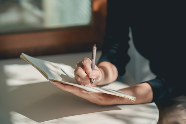 Travailleuse asiatique portant une chemise noire et un journal d'écriture sur un petit cahier sur la table au café intérieur Notes de femme et boire du café au café Concept de travail de n'importe où